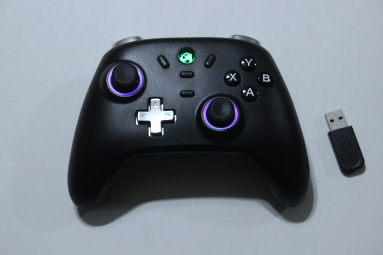 Black gaming controller with purple-lit analog sticks, D-pad, and X, Y, A, B buttons; USB dongle nearby.