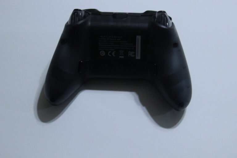 Rear view of a black game controller with textured grips resting on a white surface, showing the back and shoulder buttons