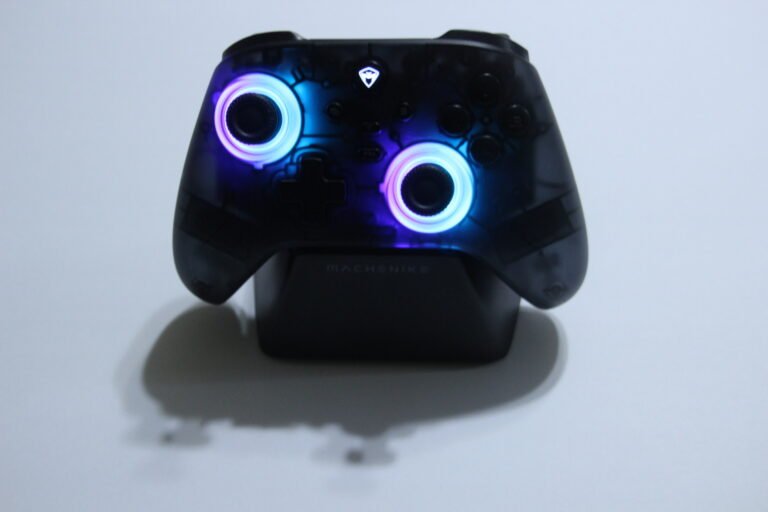 Black wireless game controller with lit blue and purple joystick rings, resting on a white surface.