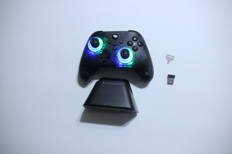 Black Machenike wireless game controller on a charging stand with blue-green RGB glow, USB dongle and SIM-ejector tool nearby