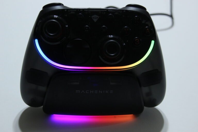Black game controller with dual analog sticks and illuminated rainbow LED arc along bottom edge, brand name MACHENIKE visible at the back.