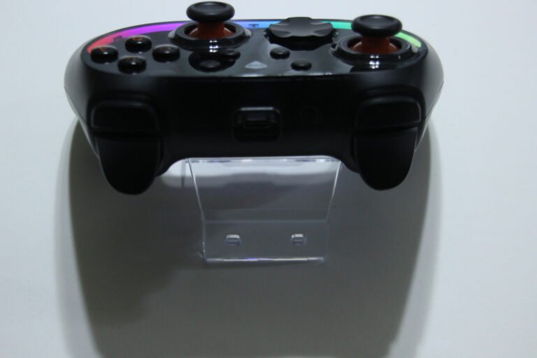 Front view of a black game controller on a clear stand, showing two analog sticks and colorful top edge bands.