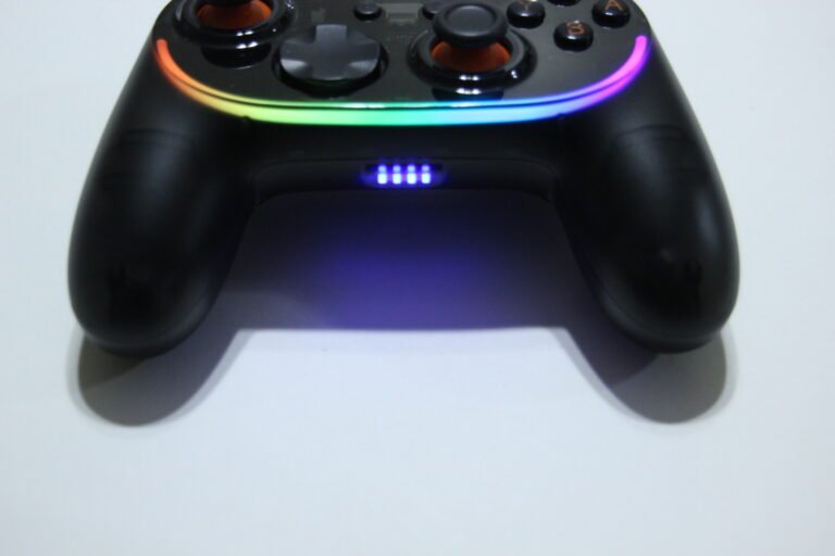 Black game controller with rainbow RGB light ring along its top edge, resting on a white surface.