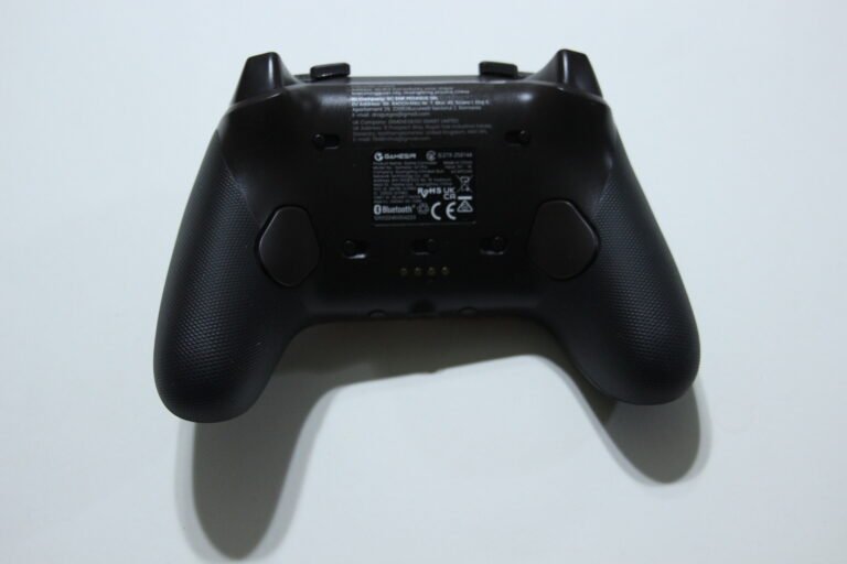 Back view of a black game controller showing a label with regulatory text on the middle back panel, on a white surface.