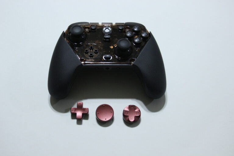 Close-up of a black Xbox-style game controller with pink detachable thumbsticks and D-pad pieces laid out on a light surface front and center on a white background.