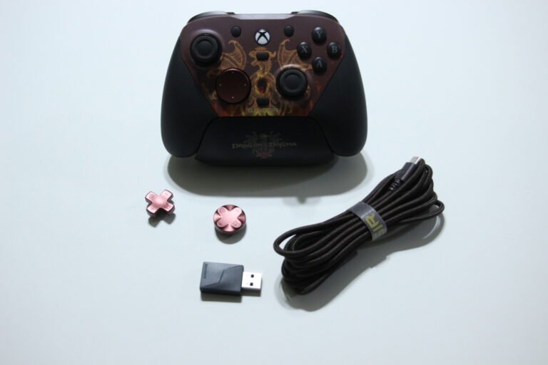 Xbox wireless controller with Dragon's Dogma design and pink accessory caps, plus a braided USB cable and small pink cross buttons on a light background